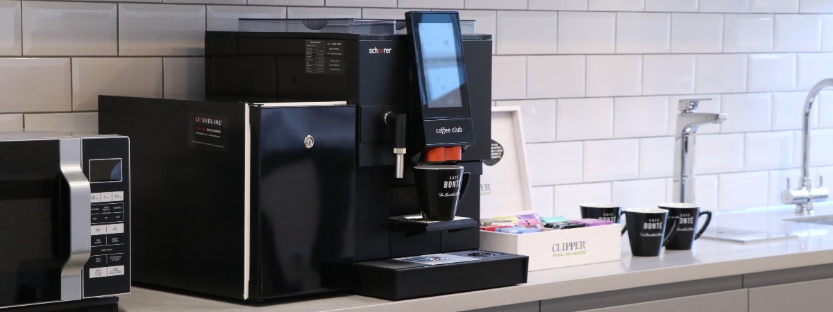 Coffee machine in sleek modern kitchen. Features white tile backsplash, microwave, mugs, and accessory box.