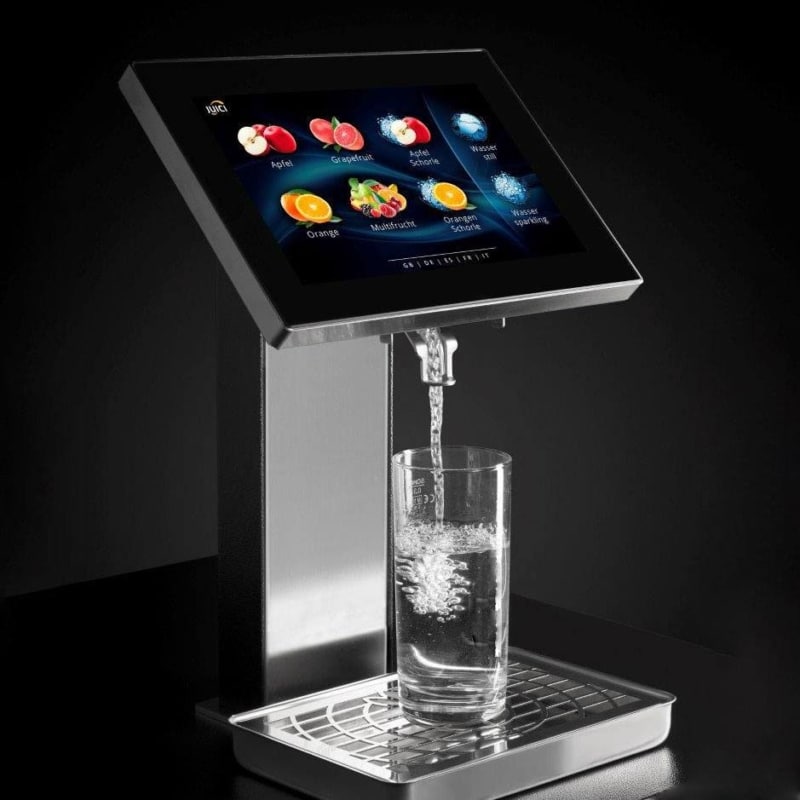 machine with a touchscreen displaying drink options like apple, grapefruit, and water. A glass sits on the metal drip tray below.