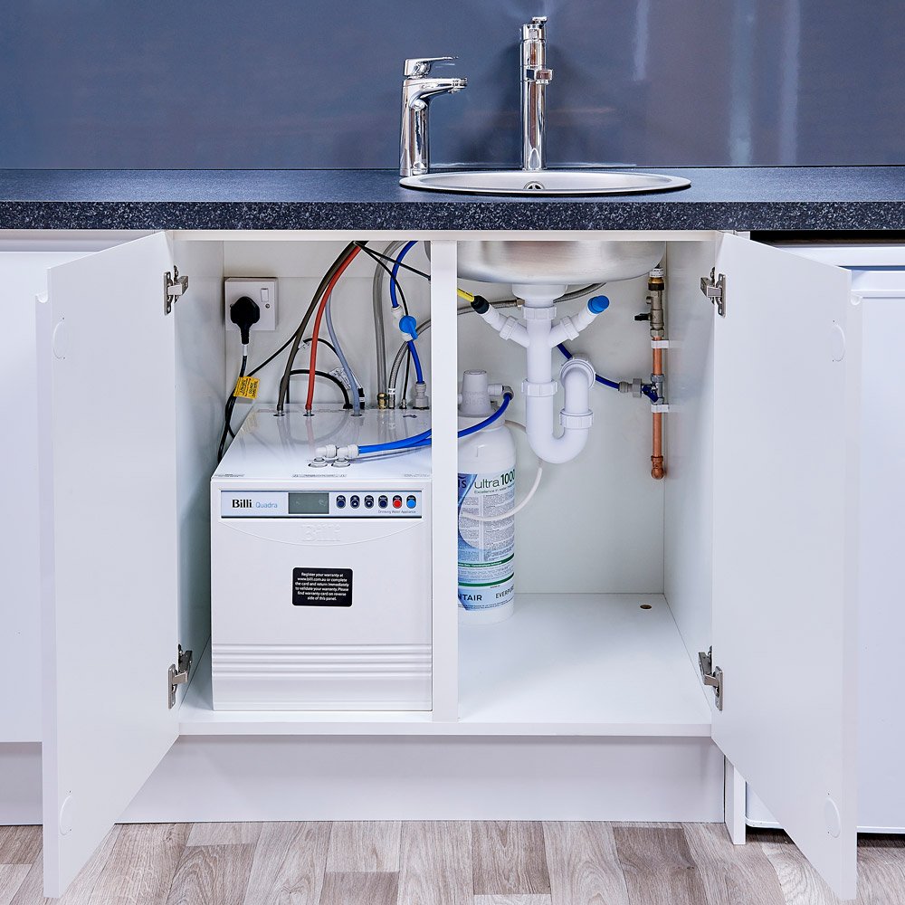 Under-sink view displaying an open cabinet with water filtration system, pipes, and cords.