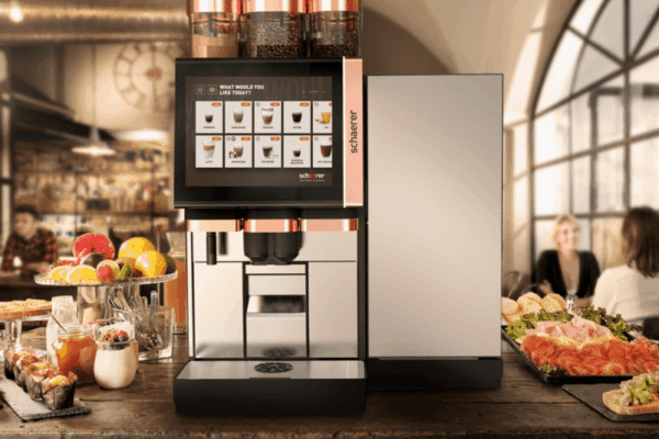 Schaerer Soul 10 6 machine with digital display on a rustic wooden counter, surrounded by fresh fruits and pastries. Cozy café setting with people chatting.