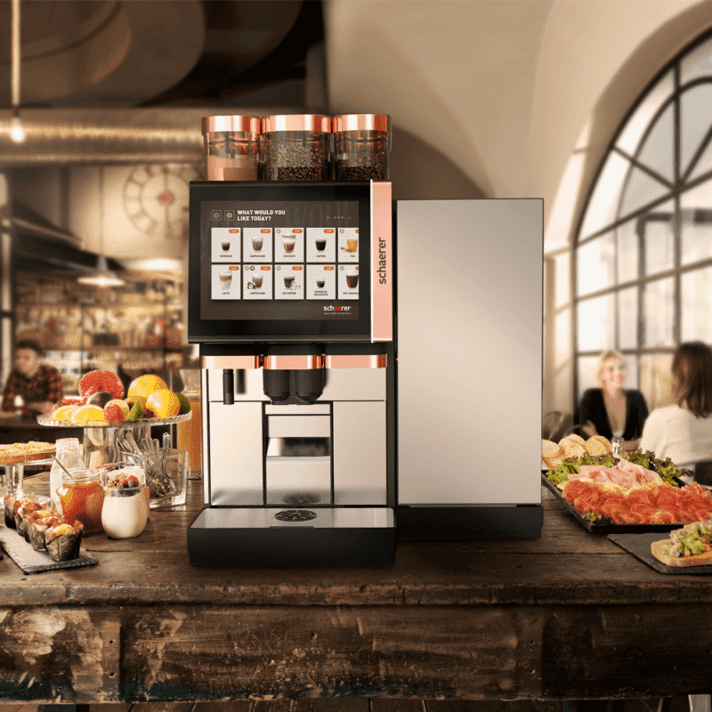 Schaerer Soul 10 6 machine with digital display on a rustic wooden counter, surrounded by fresh fruits and pastries. Cozy café setting with people chatting.